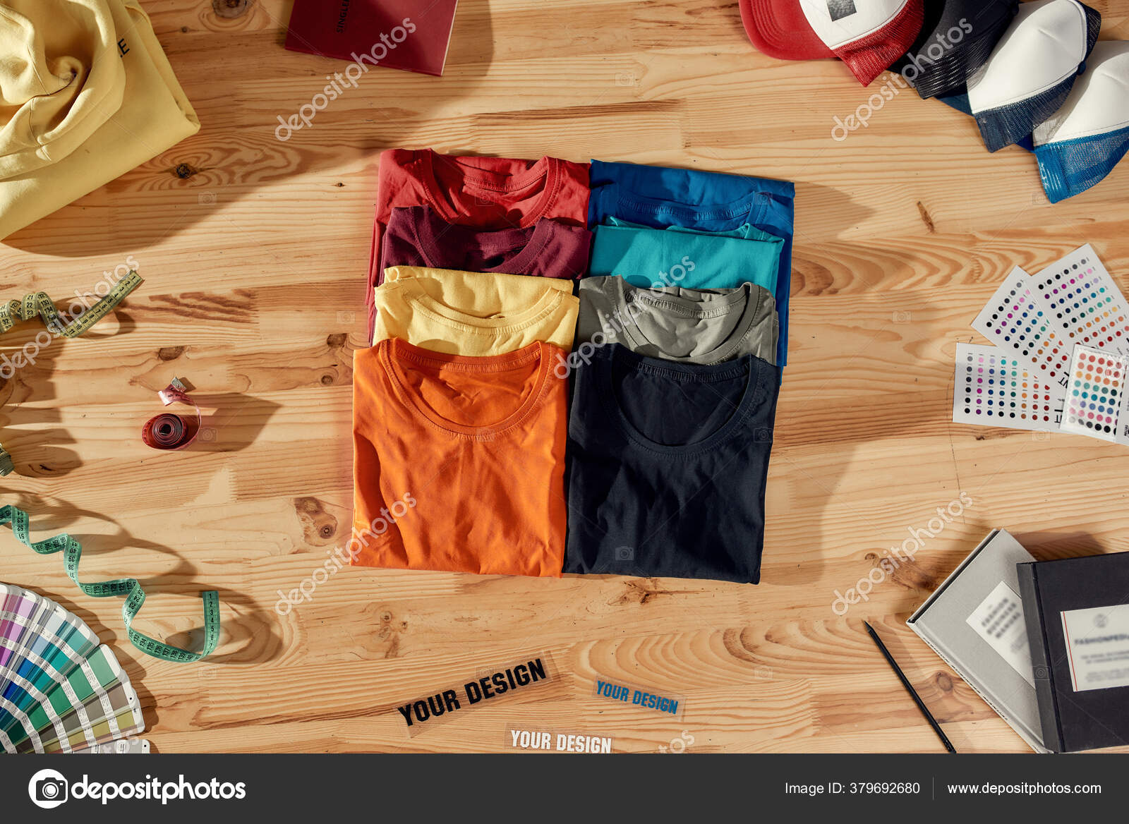 Workspace for Designer. Top view of neatly folded custom t shirts and  arranged baseball caps. Samples of fabric, stickers with text, tape measure  and notebooks lying on a wooden background. Stock Photo